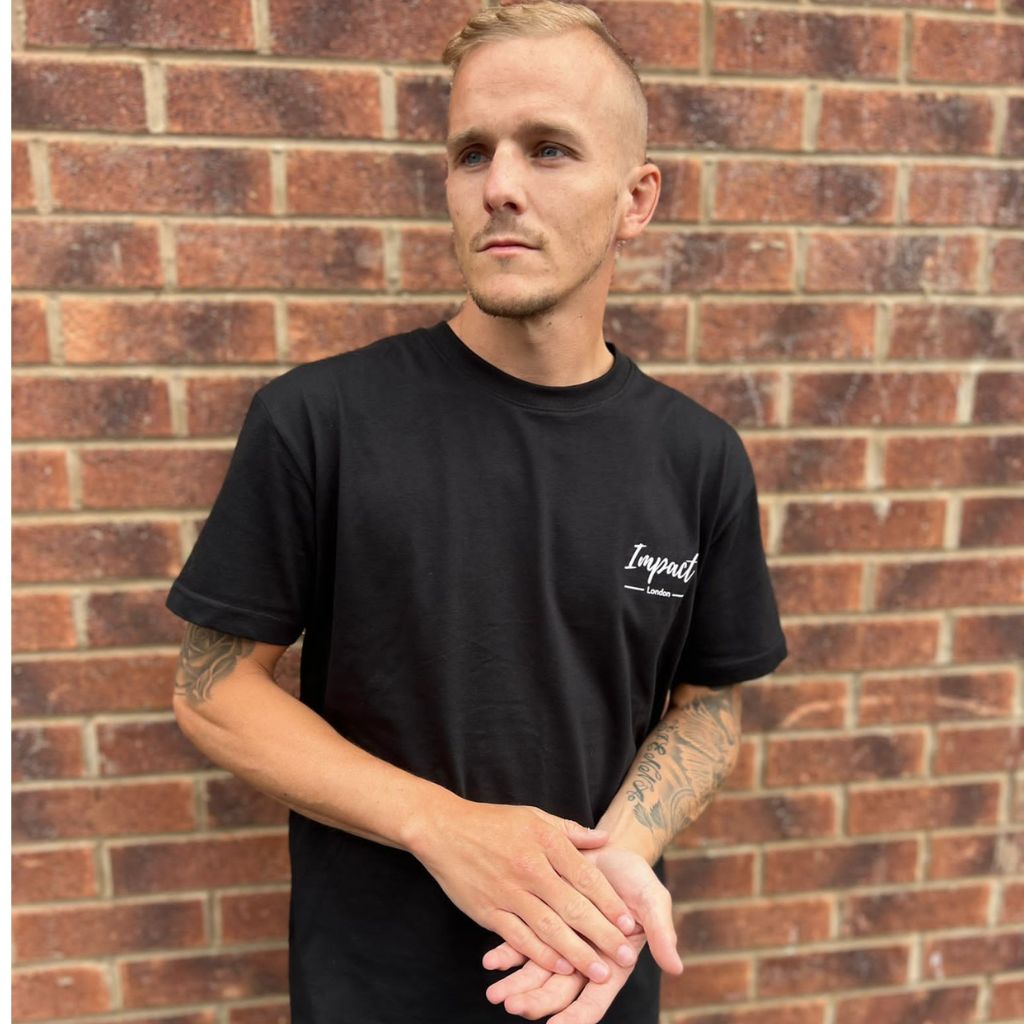 Model Liam Franklin wearing the black Impact London unisex t-shirt standing against a brick wall outside